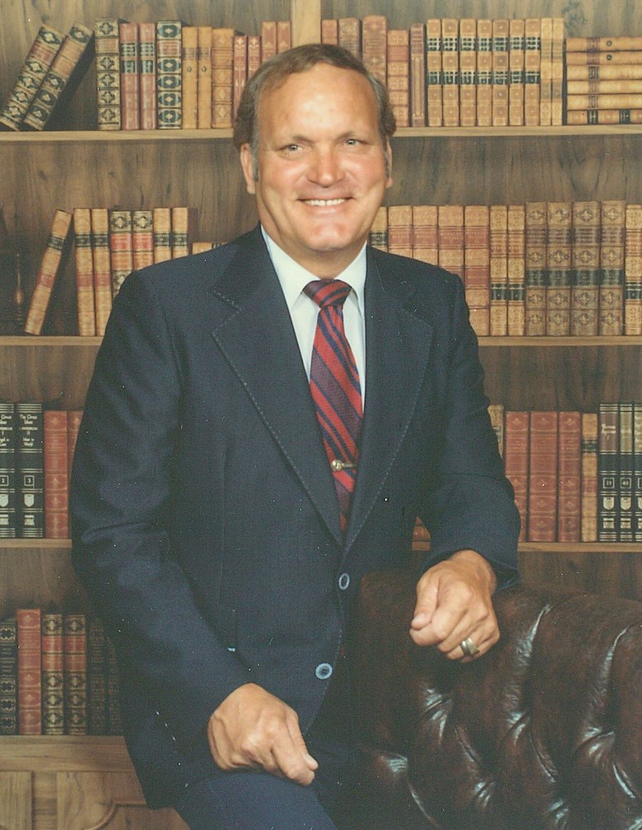 Dr. Herb Adams in a suit and tie, seated in his study in front of shelves of books